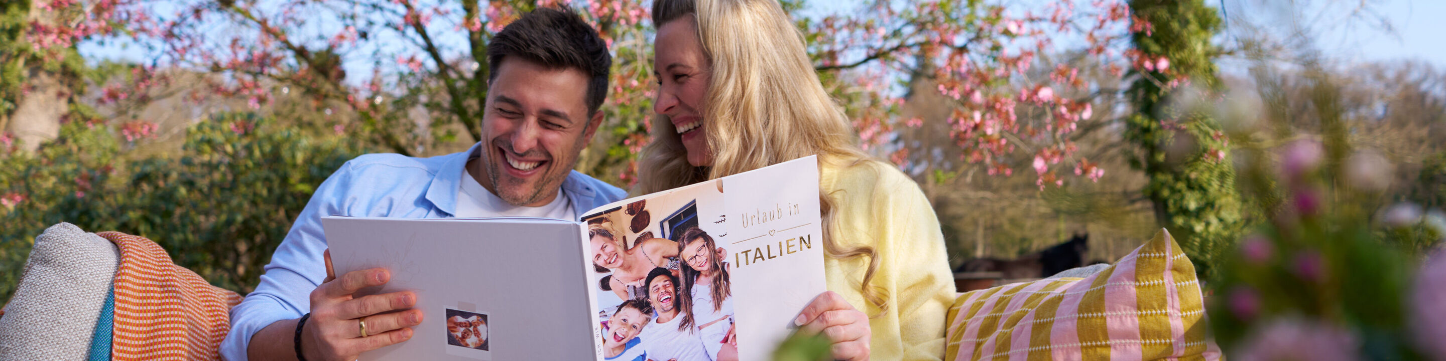 Ein lachendes Paar sitzt im blühenden Garten und betrachtet gemeinsam ein CEWE FOTOBUCH.