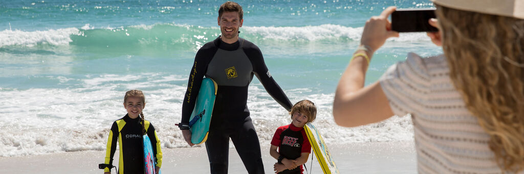 Zu sehen ist eine Familie am Strand. Eine Frau steht mit dem Rücken zur Kamera und fotografiert mit einem Smartphone ihren Mann und zwei Kinder, die mit Surfbrettern aus der Brandung kommen.