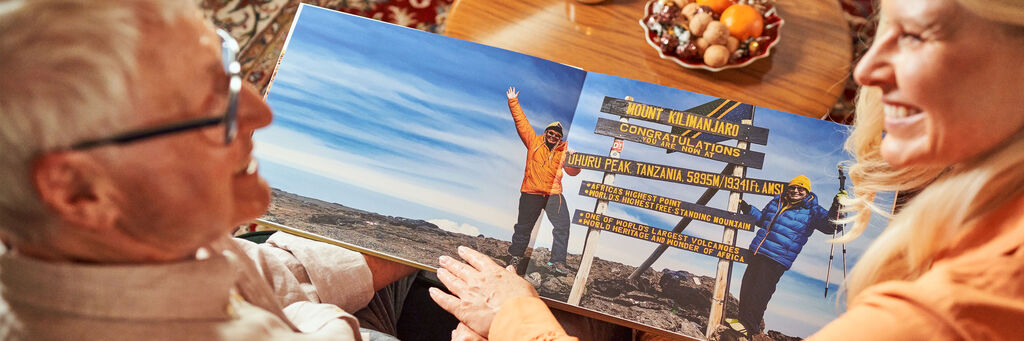 Jessica und Hans schauen sich gemeinsam ein aufgeschlagenes CEWE FOTOBUCH an und lachen sich an. Der Blick fällt über die Schulter von Jessica und Hans auf ein aufgeschlagenes CEWE FOTOBUCH, auf dem die beiden beim Posen auf dem Kilimanjaro zu sehen sind.