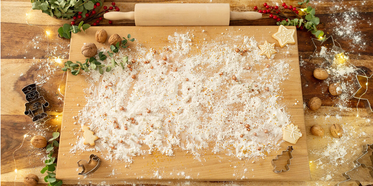 Auf dem Foto ist eine Arbeitsplatte aus Holz zu sehen, die weihnachtlich mit Zweigen, Lichterketten und Nüssen dekoriert wurde. Darauf liegt ein großes, mit Mehl bestreutes Holzbrett. Am Rand des Brettes sind Ausstechformen, Kekse und Nüsse zu erkennen.