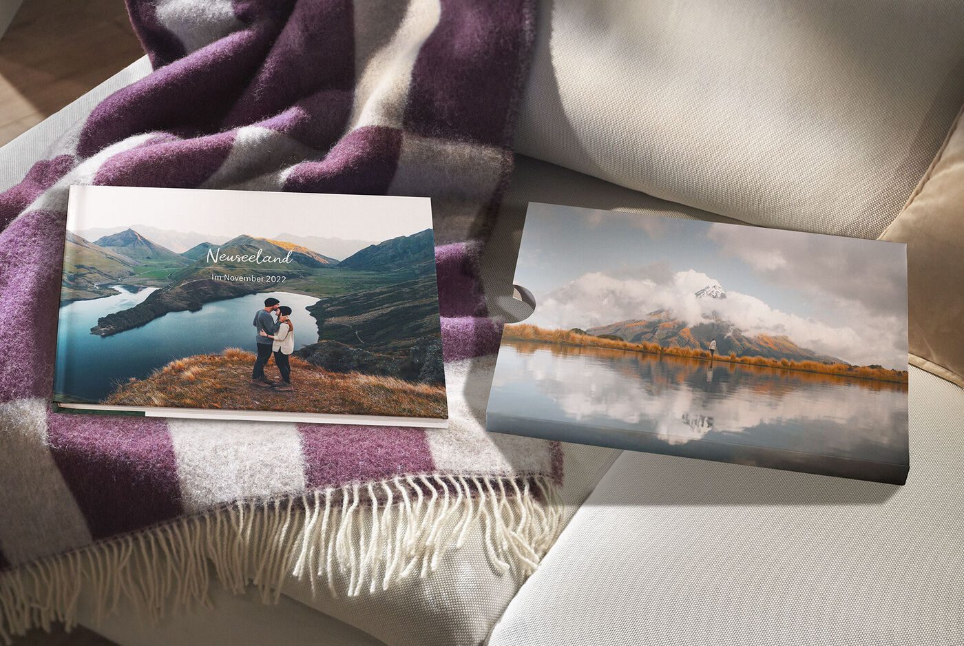 Ein CEWE FOTOBUCH liegt mit dem passenden Schuber auf einem Sofa neben einer Decke. Zu sehen sind Landschaftsmotive aus Neuseeland.