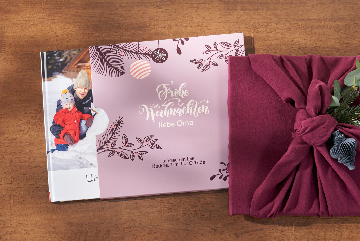 Ein CEWE FOTOBUCH mit dem Titel "Frohe Weihnachten" auf einem Holztisch. Daneben liegt ein liebevoll eingepacktes Geschenk mit einer roten Schleife.