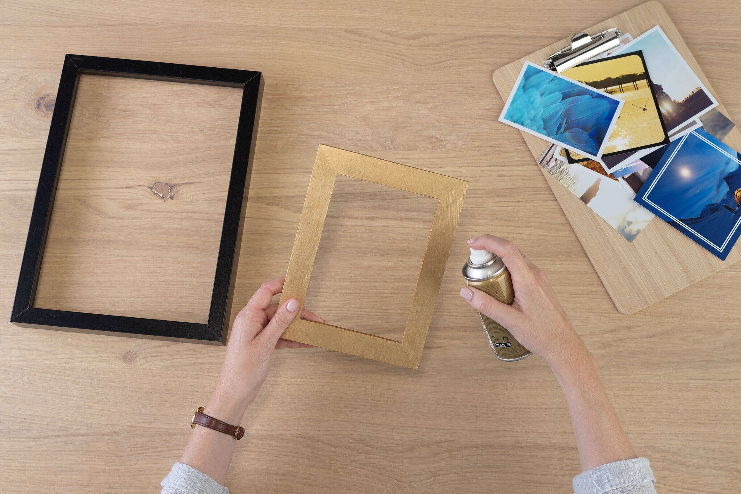 Zwei Hände halten einen Bilderrahmen und besprühen diesen mit Goldspray. Darunter liegen auf einem Tisch ein weiterer Rahmen und verschiedene Fotos.